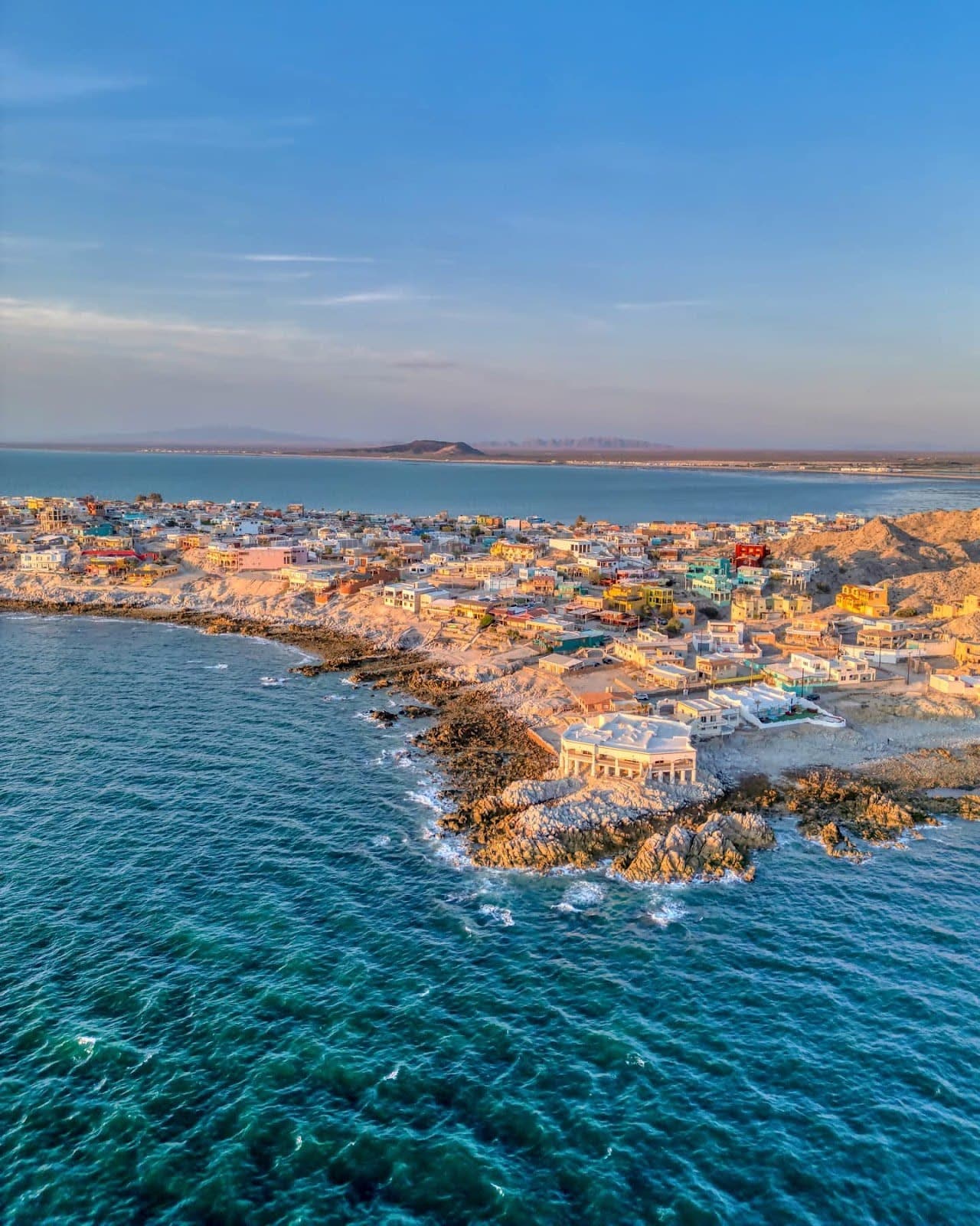 Rocky intertidal pools exposed at low tide on a desert bay in the northern Sea of Cortez, with volcanic rock formations, shallow clear water, and distant blue sea under morning light